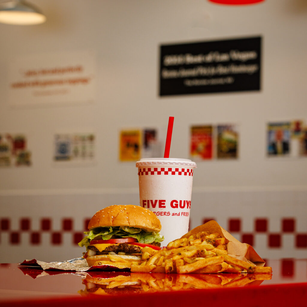 A Five Guys Little Cheeseburger, a small fry served in the new fry bag, and a drink are displayed on the iconic Five Guys counter, featuring red and white tile in the background.